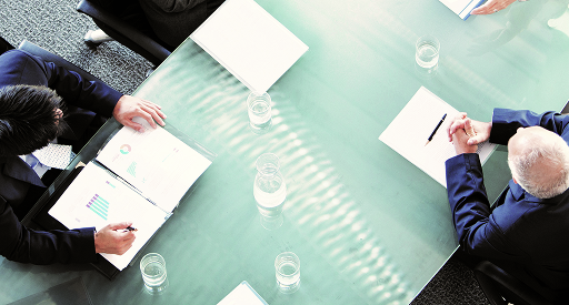 An overhead view of a meeting room with professionals holding papers seated around a glass table.