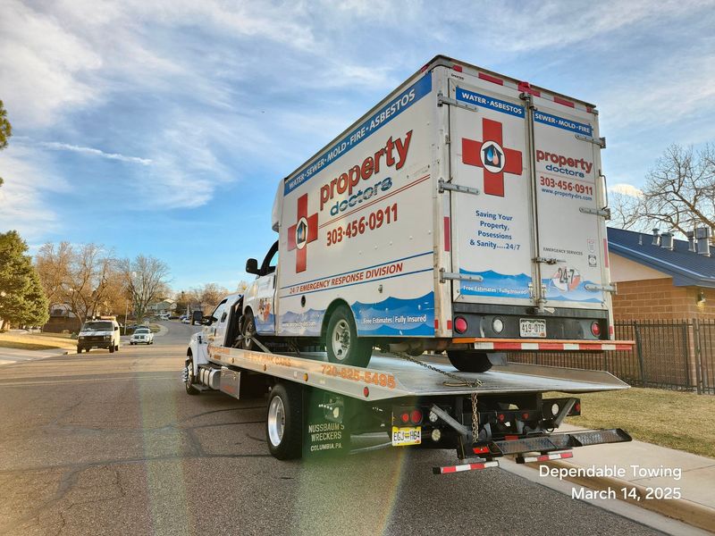 A flatbed tow truck transports a Property Doctors emergency response vehicle on a suburban road, highlighting professional towing services and reliable vehicle transport under clear daylight conditions.