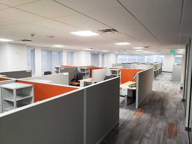 A modern office space featuring a grid of cubicles with gray walls and bright orange accent panels. Each cubicle is equipped with desks, shelving, and office chairs, providing a functional and organized workspace for employees. The space is illuminated by overhead lighting and natural light streaming in through large windows along the walls. The flooring features a contemporary gray and orange pattern, complementing the overall design.