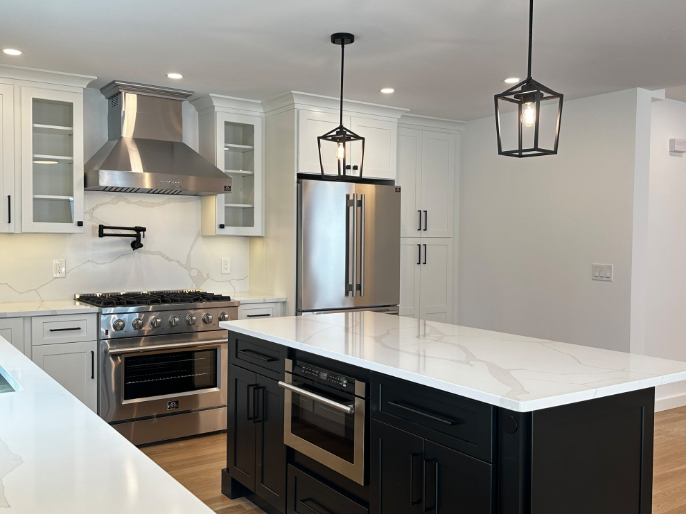 A modern, spacious kitchen with white marble countertops, stainless steel appliances, and black and white cabinetry. A large island with a microwave is positioned in the foreground, while a professional-grade gas range and a double-door refrigerator are visible in the background.