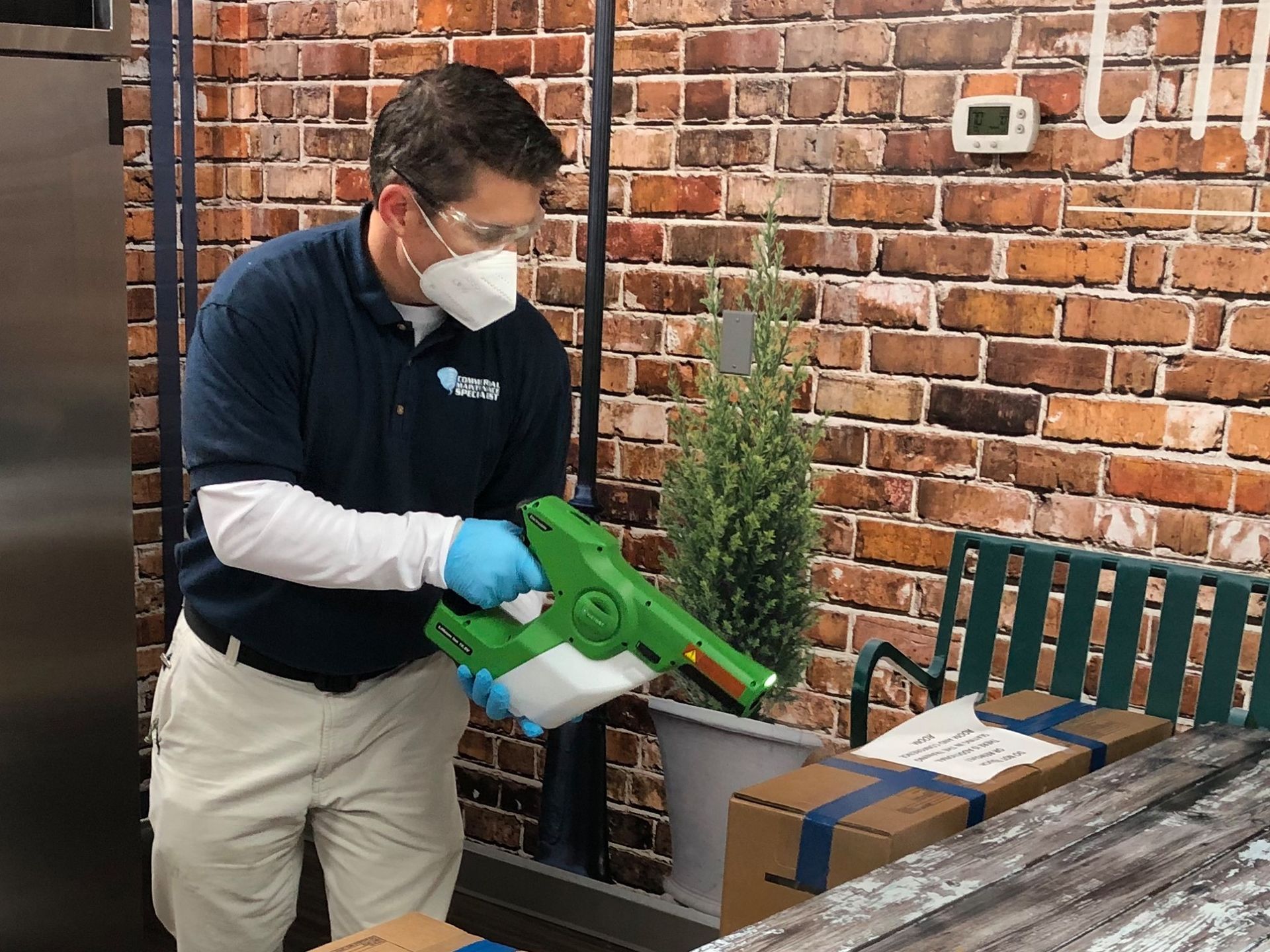 Cleaning professional wearing mask and gloves uses disinfectant sprayer to sanitize packages and surfaces in indoor workspace, highlighting health safety practices and commercial disinfection services in a maintained environment.