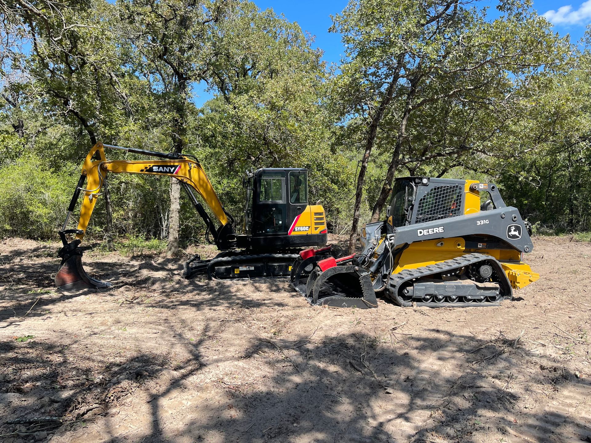 Compact excavator and skid steer clearing wooded land, removing debris and leveling soil among trees, demonstrating land clearing, brush removal, and site preparation services for residential or agricultural projects.