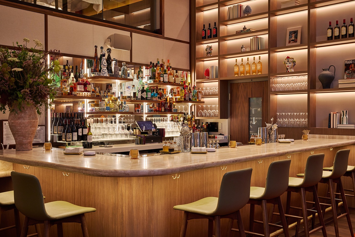 The interior of an upscale bar with warm ambient lighting, green leather bar stools, and extensive shelving filled with premium spirits and Italian wines.