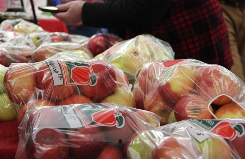Pre-packaged clear plastic bags filled with red and green locally grown apples lined up at an orchard stand. A vendor in a red plaid apron manages the produce table in the background.