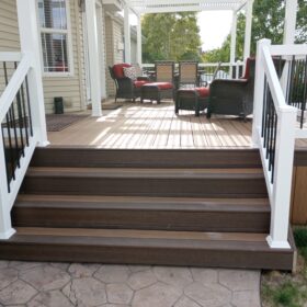 Create the perfect backyard retreat with this custom low-maintenance composite deck by DeckPro LLC. Featuring rich brown decking, a stylish white attached pergola for dappled shade, and modern white railings with black metal balusters, this outdoor space is ready for relaxing or entertaining. The wide stairs lead down to a beautiful stamped concrete patio.