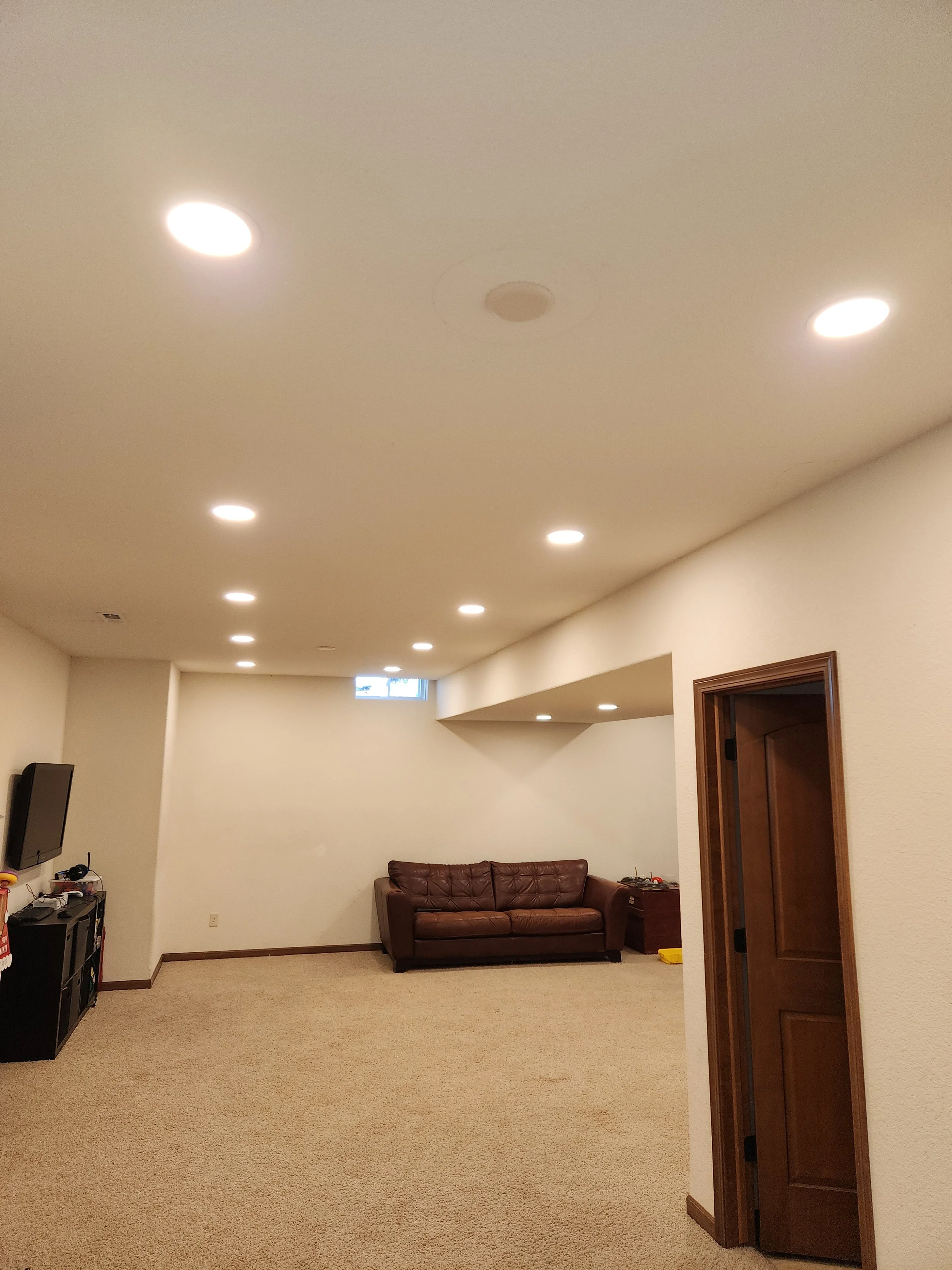 A dimly lit basement room with a brown leather couch, a television mounted on the wall, and numerous recessed lights illuminating the ceiling.