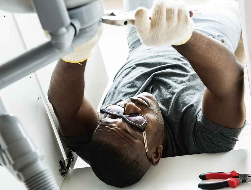 A man wearing glasses, a gray t-shirt, and light blue jeans is lying on his back, working on plumbing under a sink. He is wearing white work gloves and using a wrench to adjust a pipe. A pair of red and black pliers are visible in the foreground.