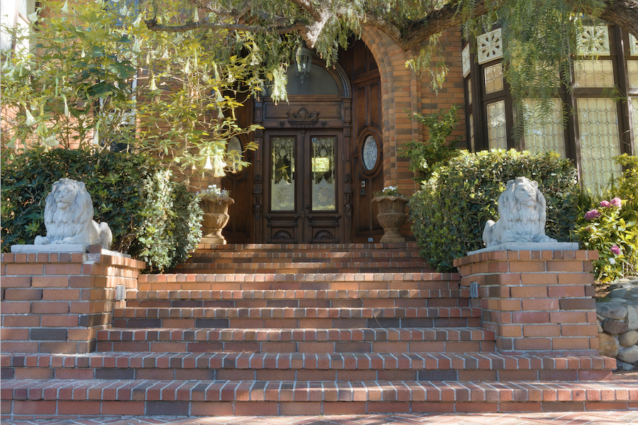Grand brick entrance with wide tiled steps, carved white lion statues, ornate wooden doors, greenery, and warm historic charm in daylight.