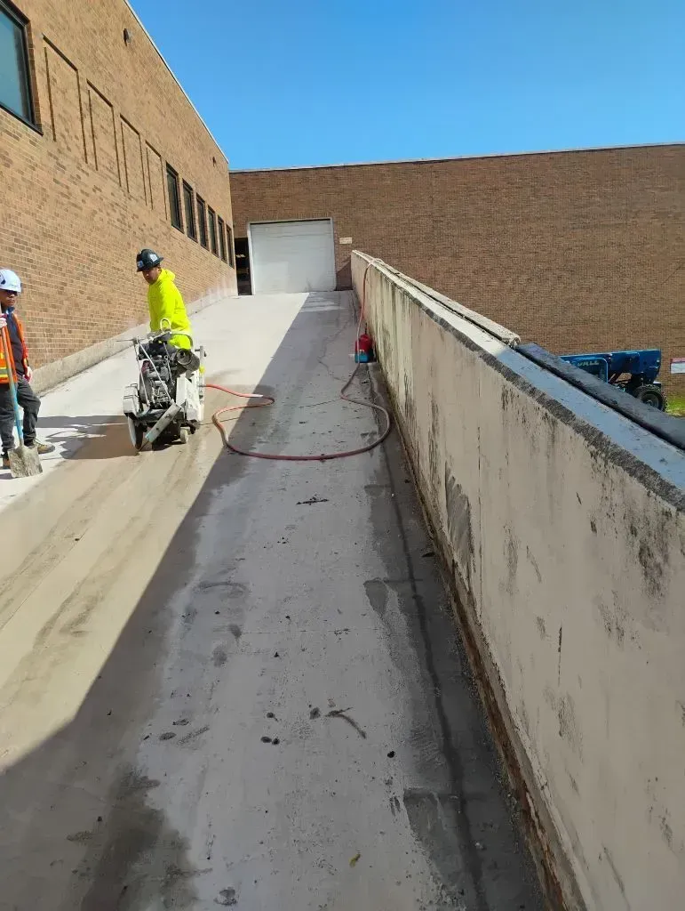Workers performing concrete cutting on sloped ramp beside commercial building, using specialized equ 