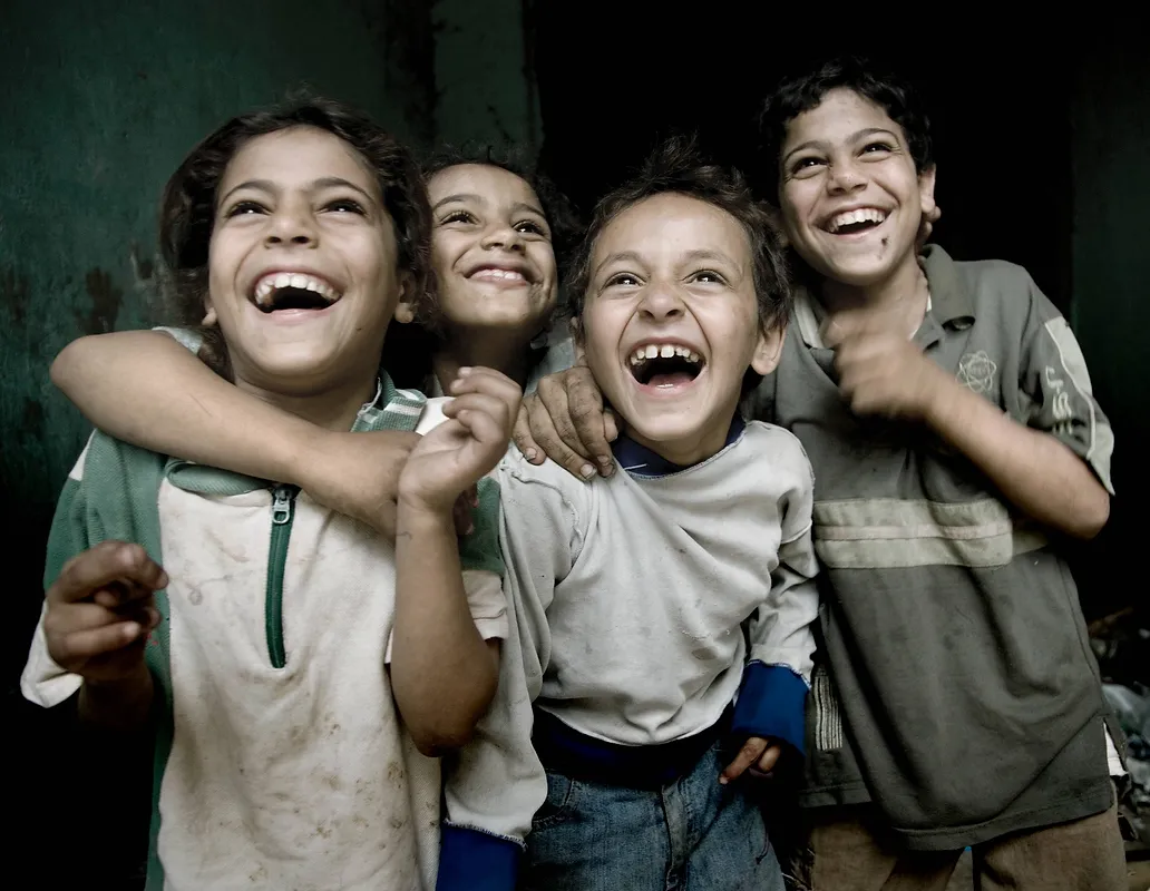 Four young children, likely brothers or close friends, are joyfully laughing with their mouths wide open. They are posing closely together, with one child's arm wrapped around another's shoulders. The children appear to be in a dimly lit, somewhat impoverished environment, with rough textures on the wall behind them. Their clothing is casual and looks worn, with some dirt smudges visible. The overall mood is one of genuine happiness and camaraderie despite their surroundings.