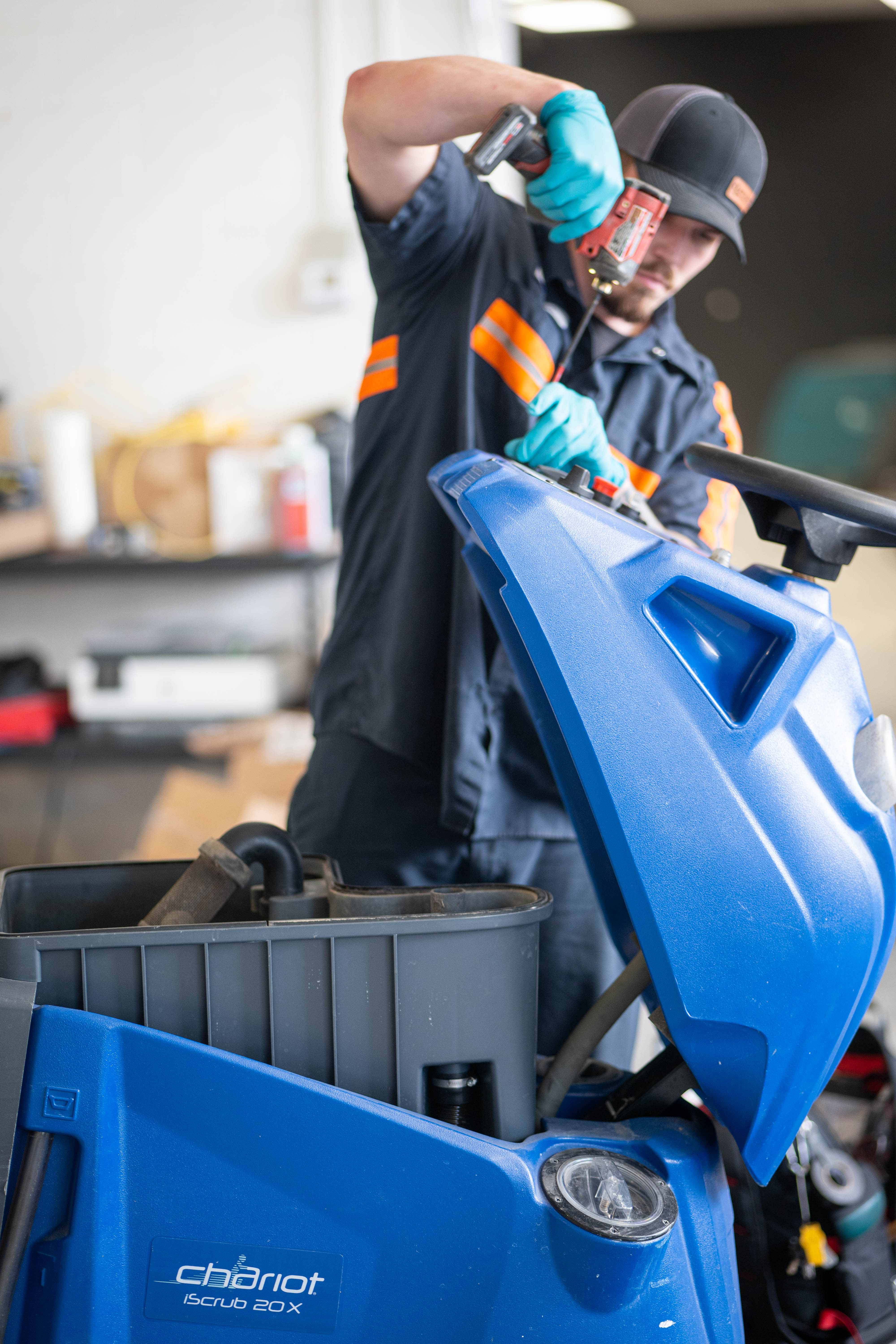 A WaySolutions service technician uses a cordless power drill to perform maintenance on the upper control console of a blue Kärcher Windsor Chariot iScrub 20X. The technician is expertly dismantling the top panel to access the internal steering and electronic components, showcasing WaySolutions' capability to service complex stand-on supermarket floor scrubbers safely and efficiently.
