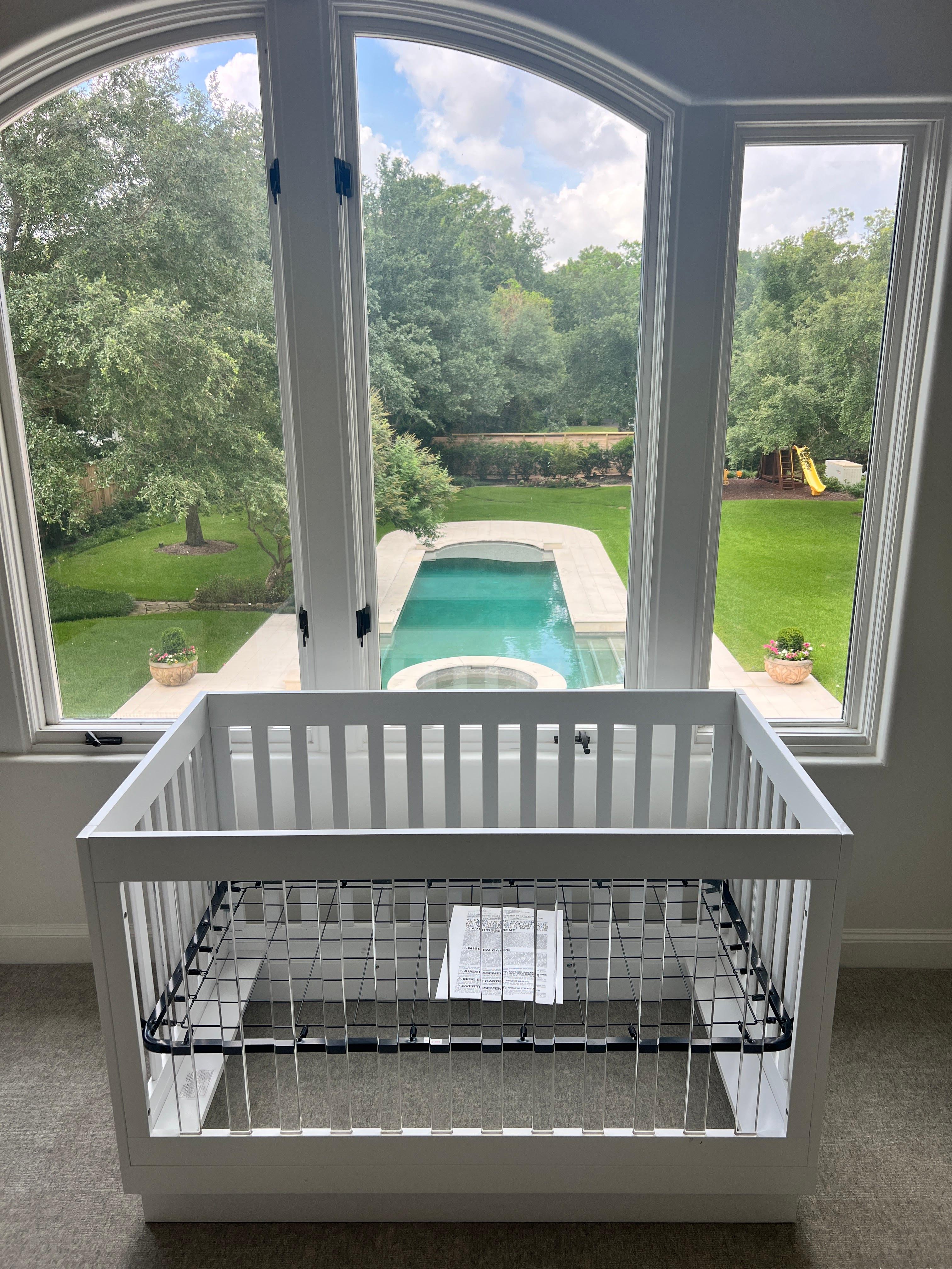 White baby crib professionally assembled in a nursery with large windows overlooking a backyard pool.