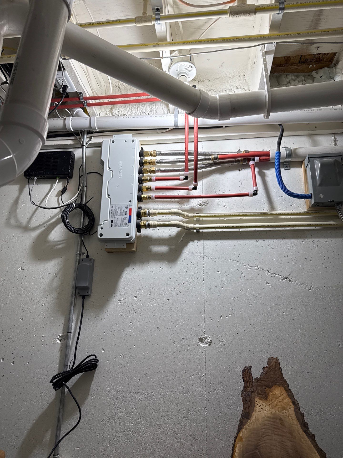 Basement plumbing manifold mounted on a wall connects multiple red and white water lines, surrounded by larger PVC drainage pipes, electrical boxes, wiring, conduit, and exposed ceiling utility components overhead.