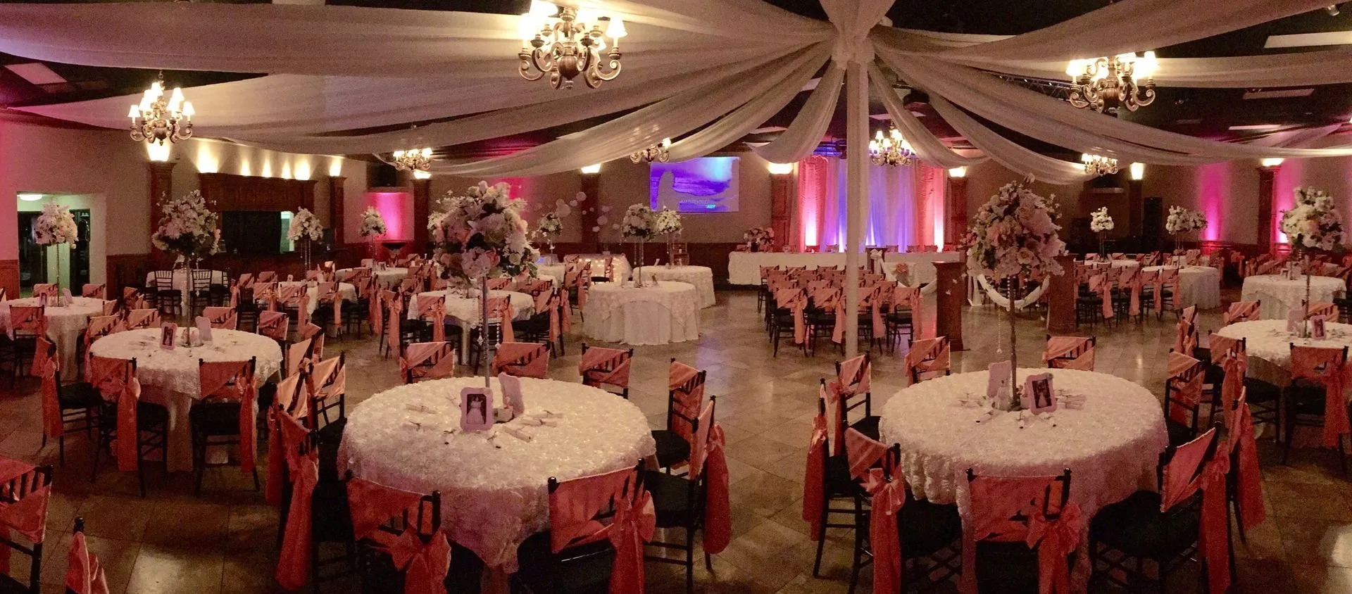 Romantic banquet hall decorated with pink lighting, floral centerpieces, ceiling drapery, round rece 