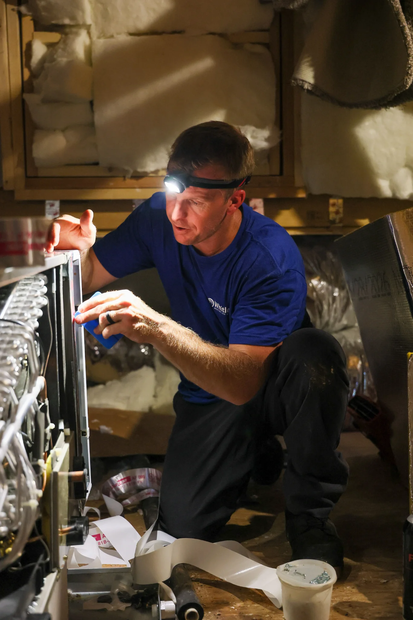 A man in a blue t-shirt and black pants, wearing a headlamp, kneels and works on an HVAC unit. He is 