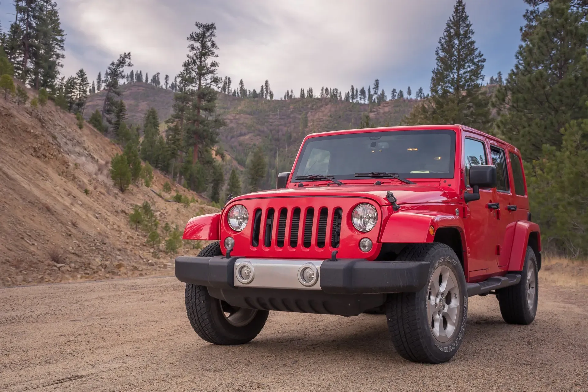 A red Jeep Wrangler is parked on a dirt road in a mountainous, forested area.