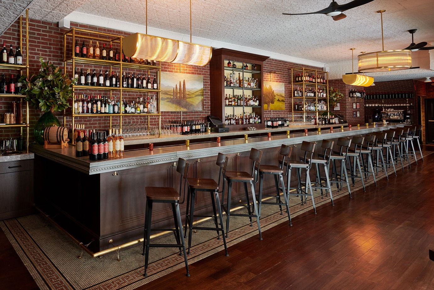 A wide shot of a wood-paneled bar with a marble top, brass shelving, and leather stools. The background features a brick wall, landscape paintings, and warm, modern light fixtures.