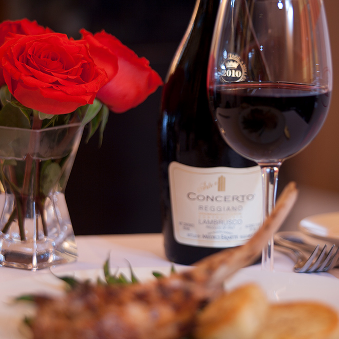 A bottle of red wine behind a glass filled with red wine with a small bouquet of red roses on a white table.
