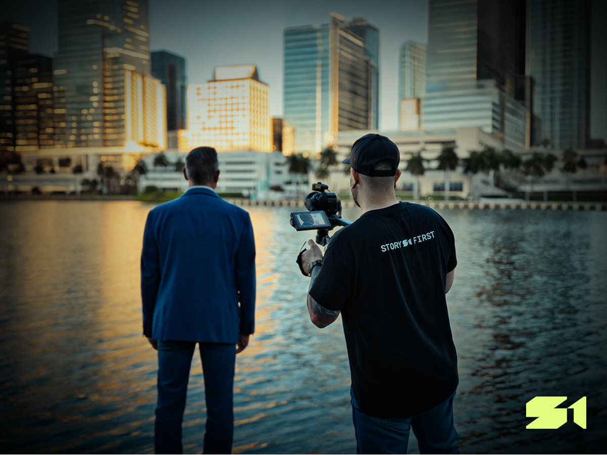 Behind-the-scenes photo of Story First filming a law firm brand video on location, with founder Chris Gray operating the camera as an attorney stands near the water in front of a city skyline.