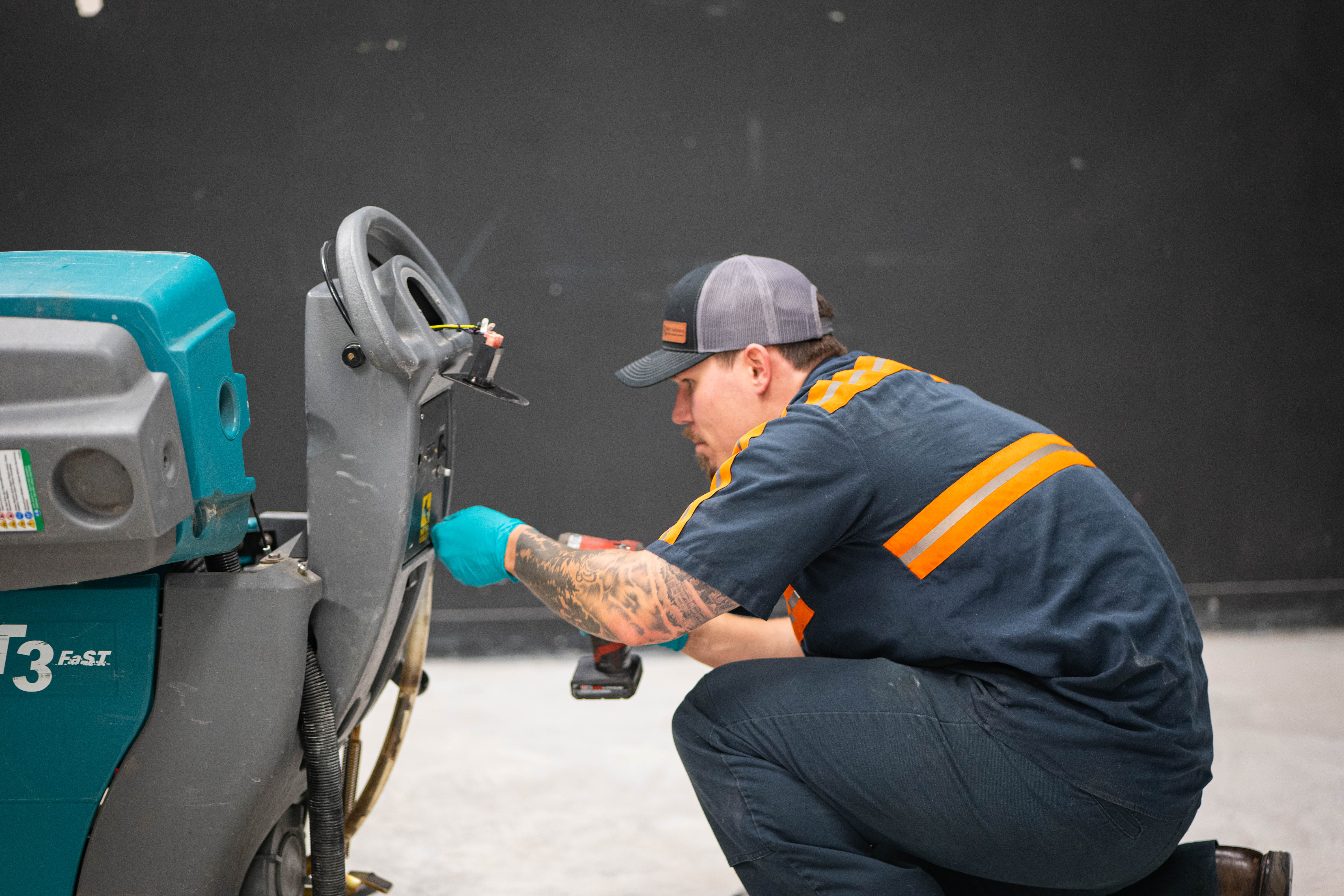 A WaySolutions field technician utilizes a power drill to safely and efficiently access the rear control panel of a teal Tennant T3 FAST walk-behind floor scrubber. Our rapid, on-site mechanical repair services in Layton, Utah, are designed to minimize equipment downtime and get your commercial cleaning fleet back to work faster.