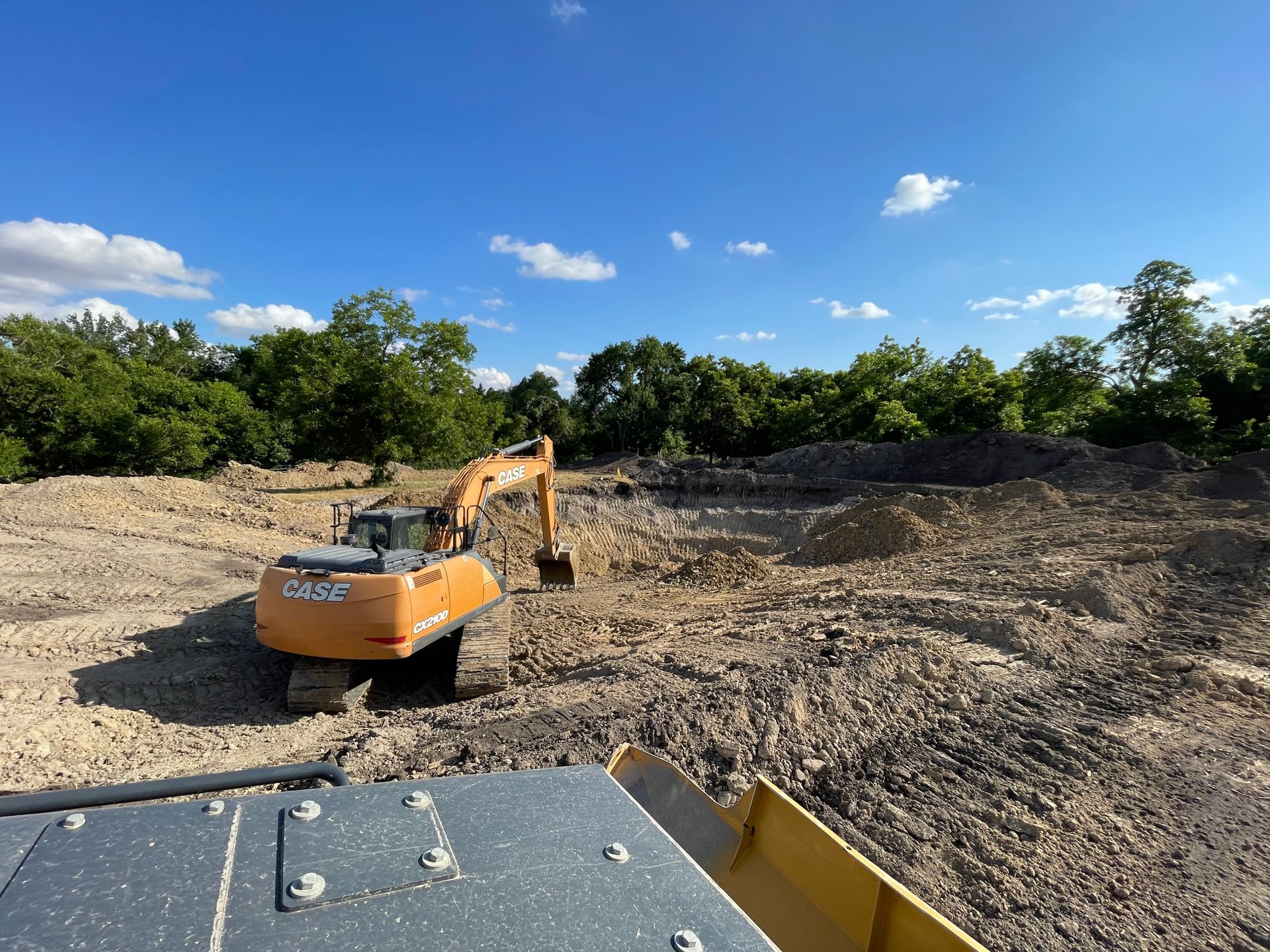Large excavator digging and grading a wide construction site under clear blue sky, surrounded by trees, showcasing earthmoving, land clearing, and site preparation work for residential or commercial development.
