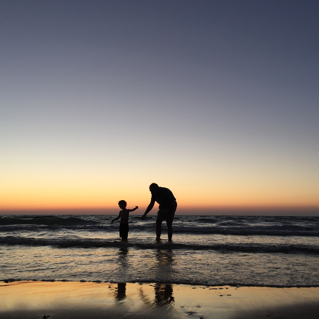 Silhouettes of adult and child standing in shallow water on the beach at sunset.