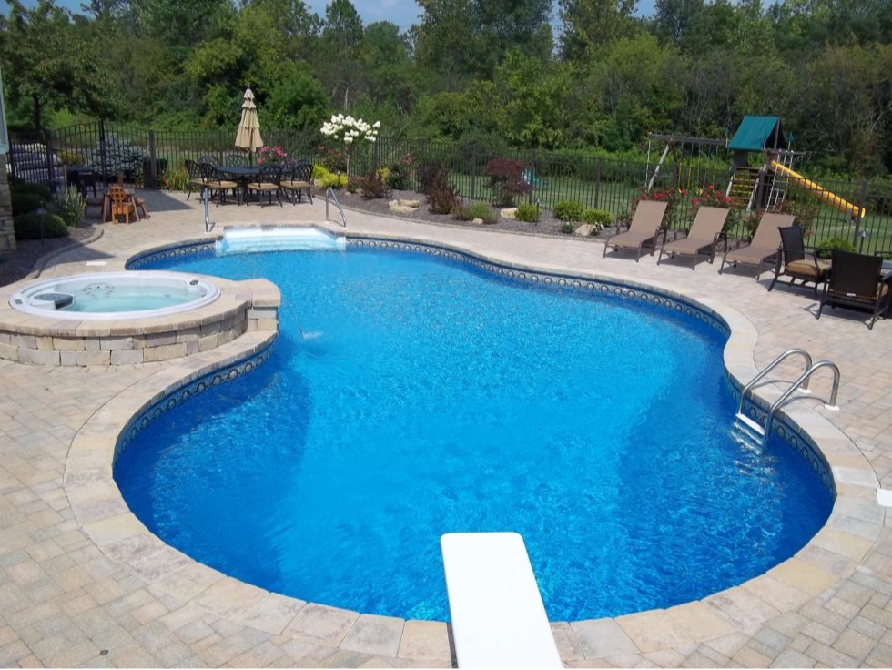 An aerial-angle view of a Latham Aspen freeform inground pool with a deep blue vinyl liner and decorative tile border is shown in a fully finished residential backyard. The pool features a white diving board at the near end and a built-in spillover spa with a raised paver surround and an adjacent round hot tub on the upper left. The expansive tan paver patio surrounds the entire pool and includes lounge chairs, a wrought iron dining set with umbrella, and lush garden beds with flowering shrubs along a black aluminum fence. A children's wooden swing set with a slide is visible in the background right, and mature trees line the property perimeter, creating a private, resort-style backyard retreat.
