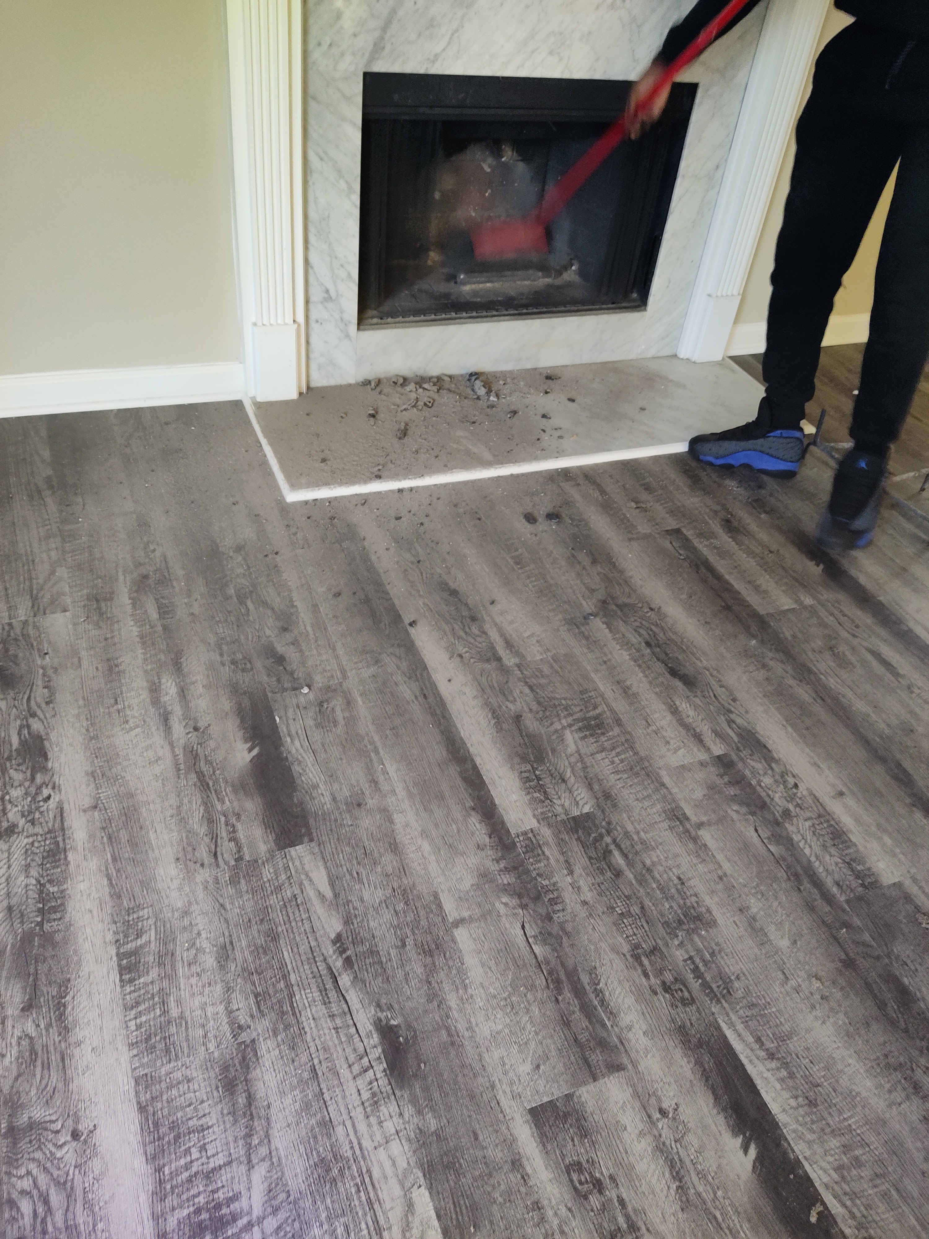 A person in black pants and sneakers is sweeping debris from a fireplace onto a grey wood-look floor. The fireplace has a marble surround and a black insert. Debris is scattered around the fireplace hearth and on the floor.