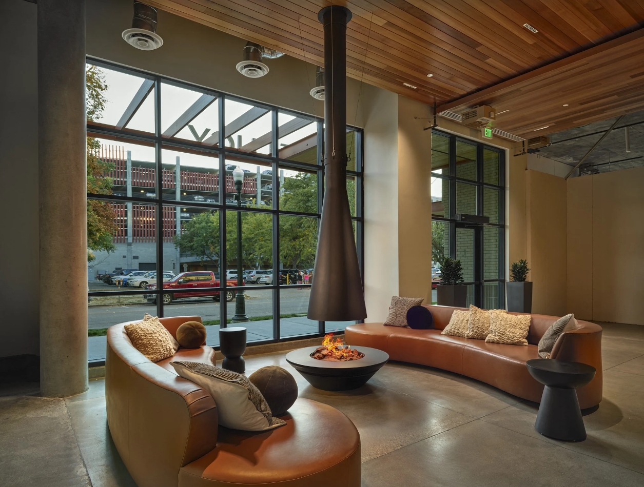 Midcentury modern resident lounge space with an indoor fire pit and chic black flame hood extending from the ceiling with large metal grid-style windows and two brown leather-style couches