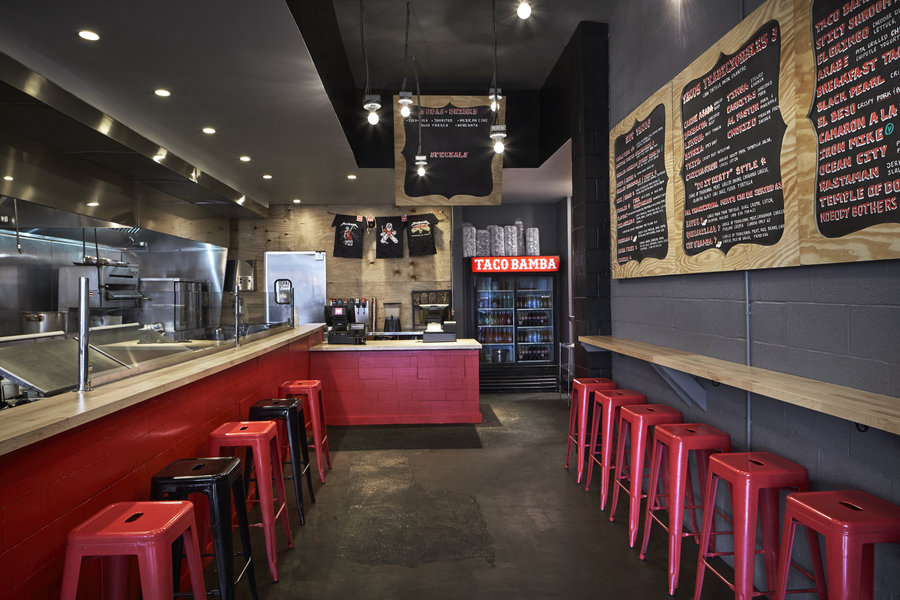 Interior of restaurant with kitchen on the left with a glass divider and a small counter with stools. another small counter and stool on the opposite wall with cash register at the back of the restaurant.