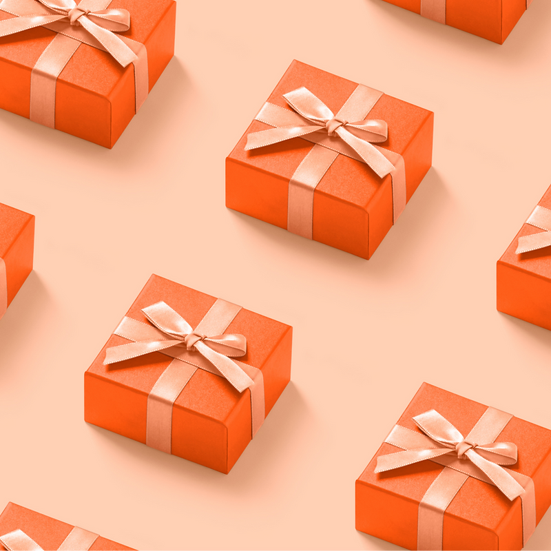 Several orange gift boxes with satin ribbons are arranged in a repeating pattern on a light orange background. The boxes are square and have a lid tied with a ribbon and a bow. The lighting creates subtle shadows, giving the boxes a three-dimensional appearance.