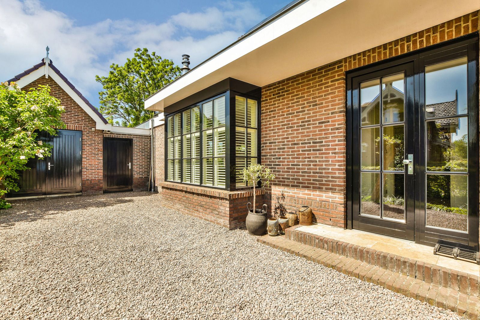 A charming brick house with large black-framed windows and a white overhang sits beside a gravel driveway. An old brick outbuilding is visible in the background.