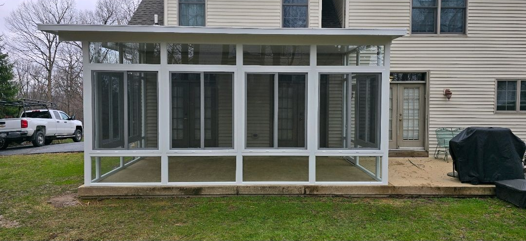 Contemporary screened-in porch attached to a residential home, featuring white aluminum framing, large mesh panels, and a flat roof. The structure creates a bright, enclosed outdoor space ideal for relaxing while enjoying airflow and protection from insects.