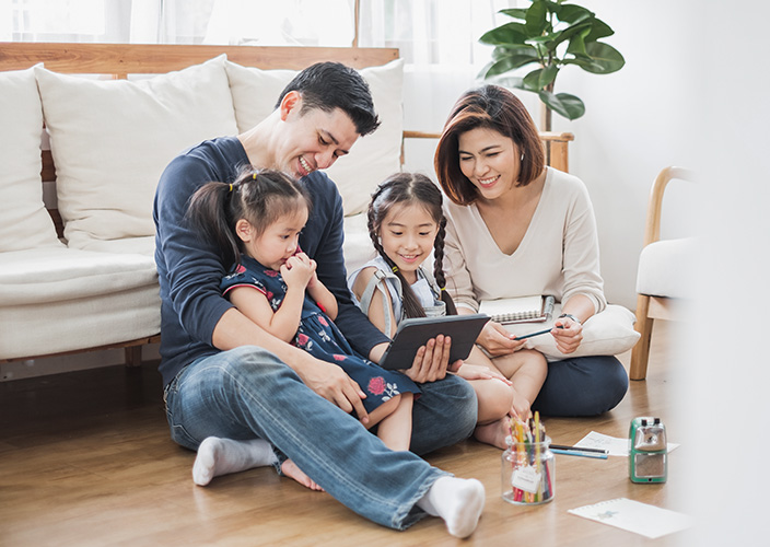 Family of four sitting on floor looking at tablet together in bright living room.