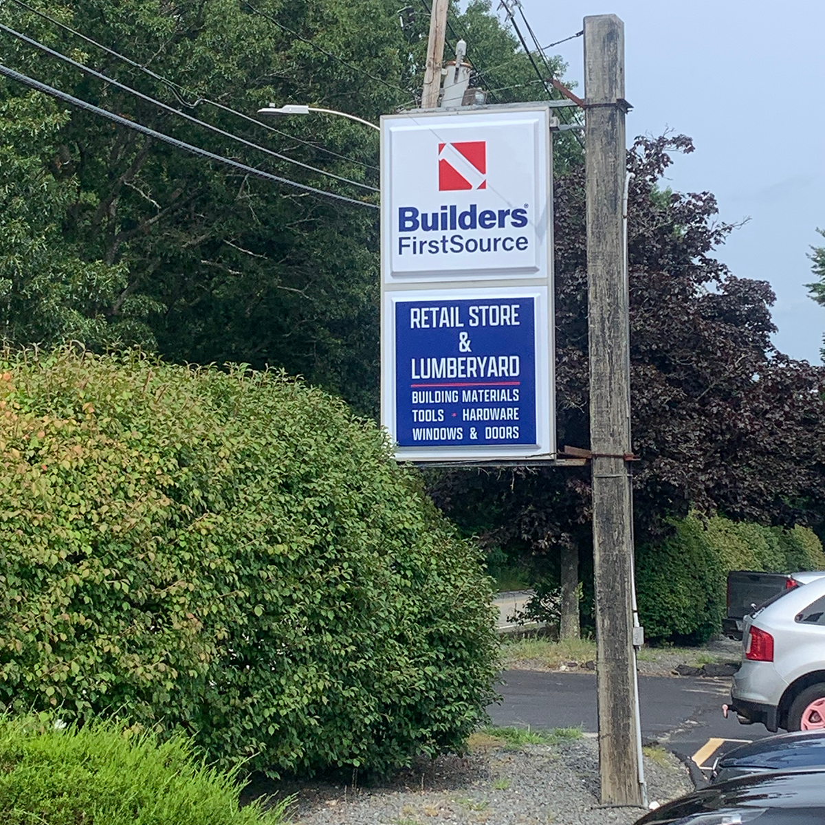 An outdoor Builders FirstSource sign stands beside a wooden utility pole, leafy trees, and landscaped shrubs. The sign’s upper panel displays the Builders FirstSource logo, while the lower blue panel reads “Retail Store & Lumberyard” and lists “Building Materials,” “Tools + Hardware,” and “Windows & Doors.” Parked vehicles are partially visible in the foreground and right side of the image.