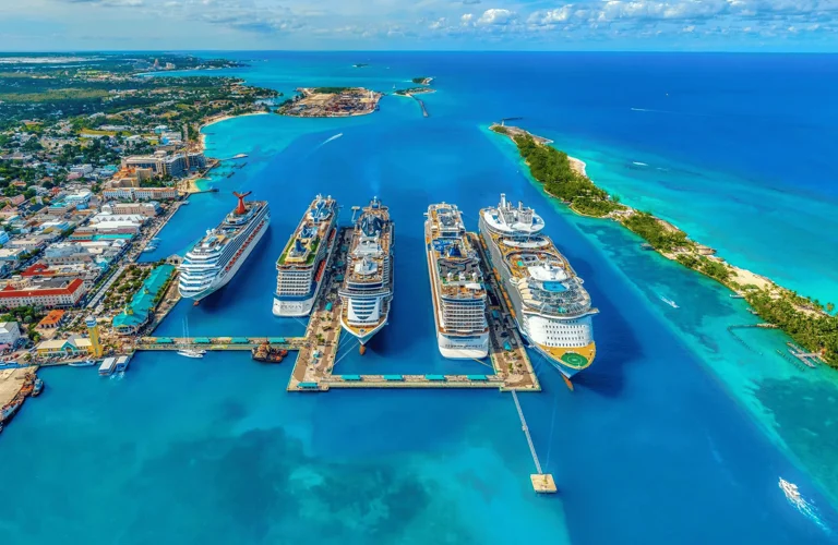 An aerial drone shot captures a bustling cruise port on a bright, sunny day, with several large cruise ships docked at a pier. The vibrant turquoise water surrounds the ships, contrasting with the lush green of the nearby island. In the background, a coastal town with colorful buildings and a lighthouse is visible, along with other islands and the vast expanse of the ocean.