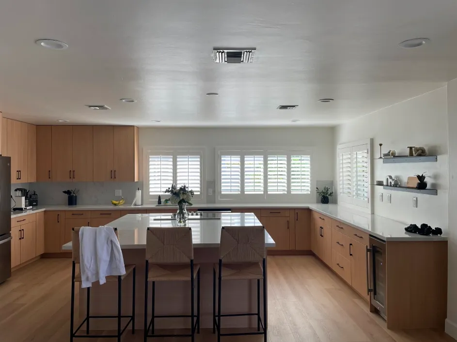 Modern open-concept kitchen with light wood cabinetry, white countertops, central island with bar stools, stainless appliances, plantation shutters, and minimalist decor creating a bright, clean, and inviting interior space overall.