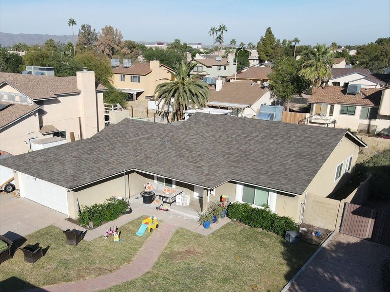 An aerial shot showcasing a suburban home with a newly installed brown asphalt shingle roof. The house features tan siding, a white garage, and a green lawn with children's toys.