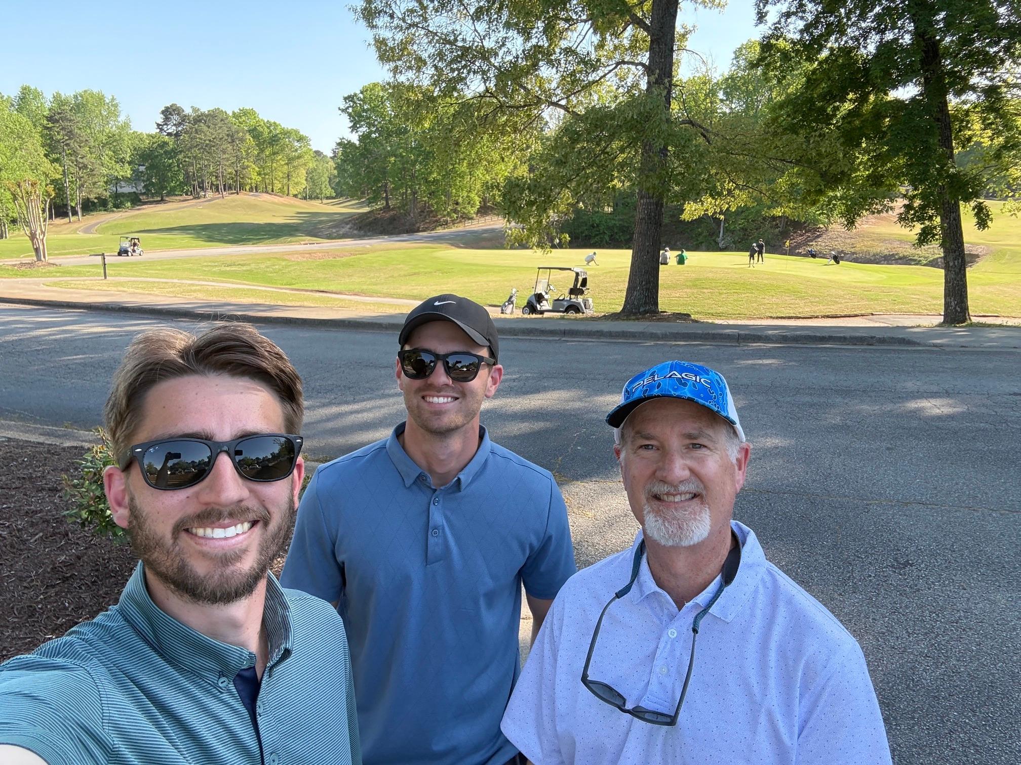 Three smiling men posing for a selfie on a sunny golf course with carts and fairway visible in the background.