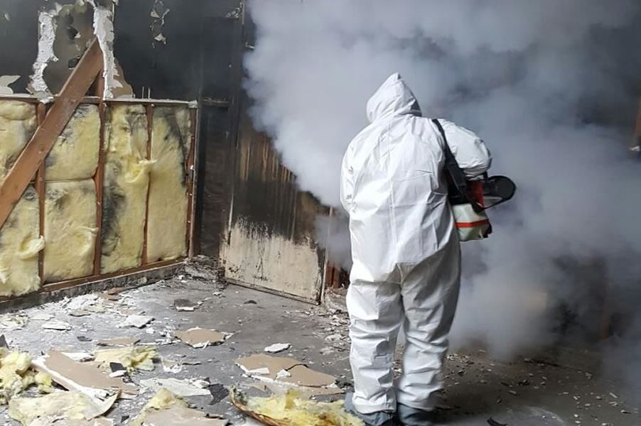 A person in a white hazmat suit stands in a room with exposed insulation, debris scattered on the floor, and smoke or fog emanating from a device they are holding. The walls show signs of damage and charring.