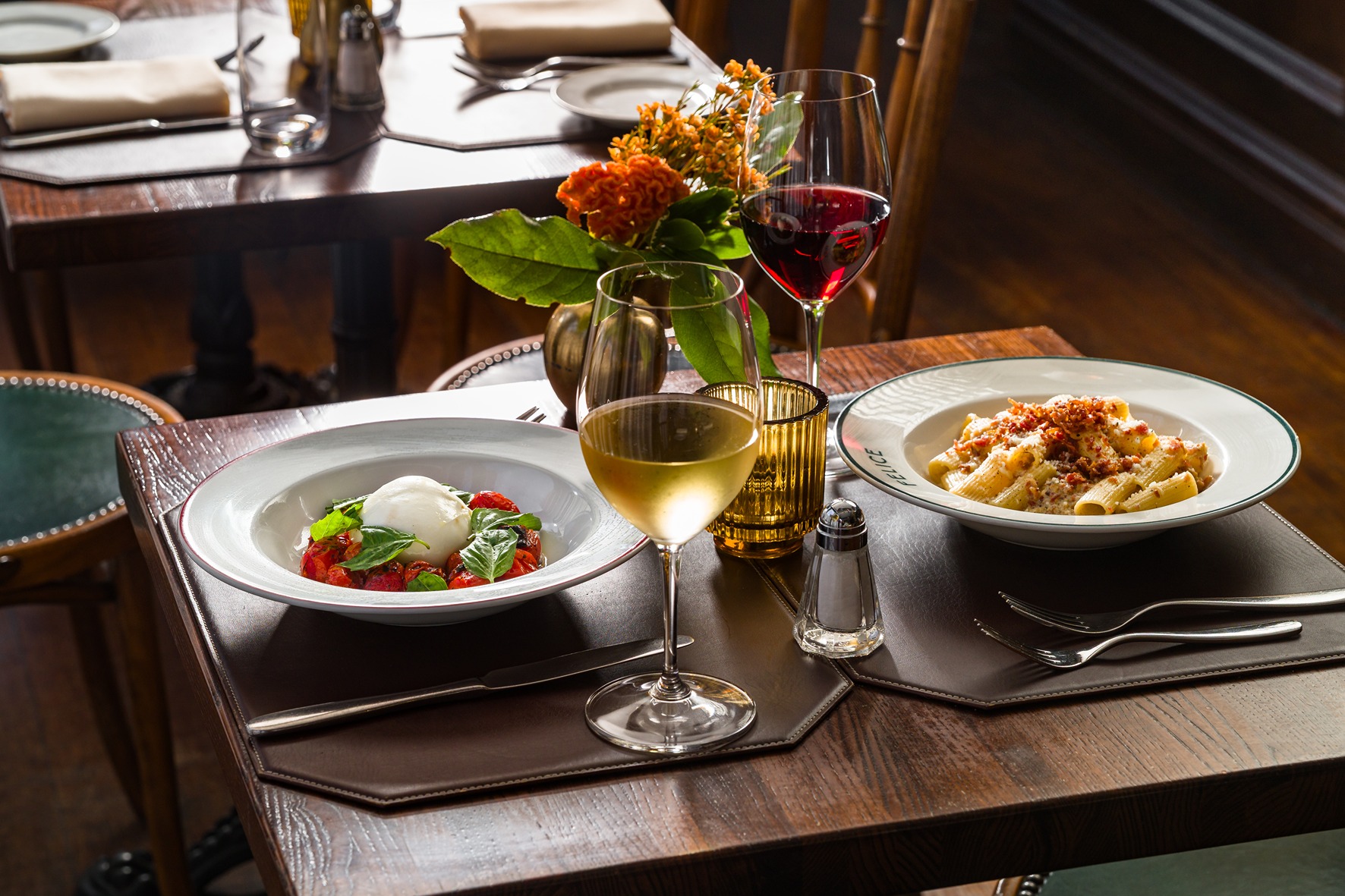 A cozy restaurant table featuring a plate of rigatoni, a burrata appetizer, and glasses of red and white wine. The scene is lit with warm, natural light near a dark wood wall.