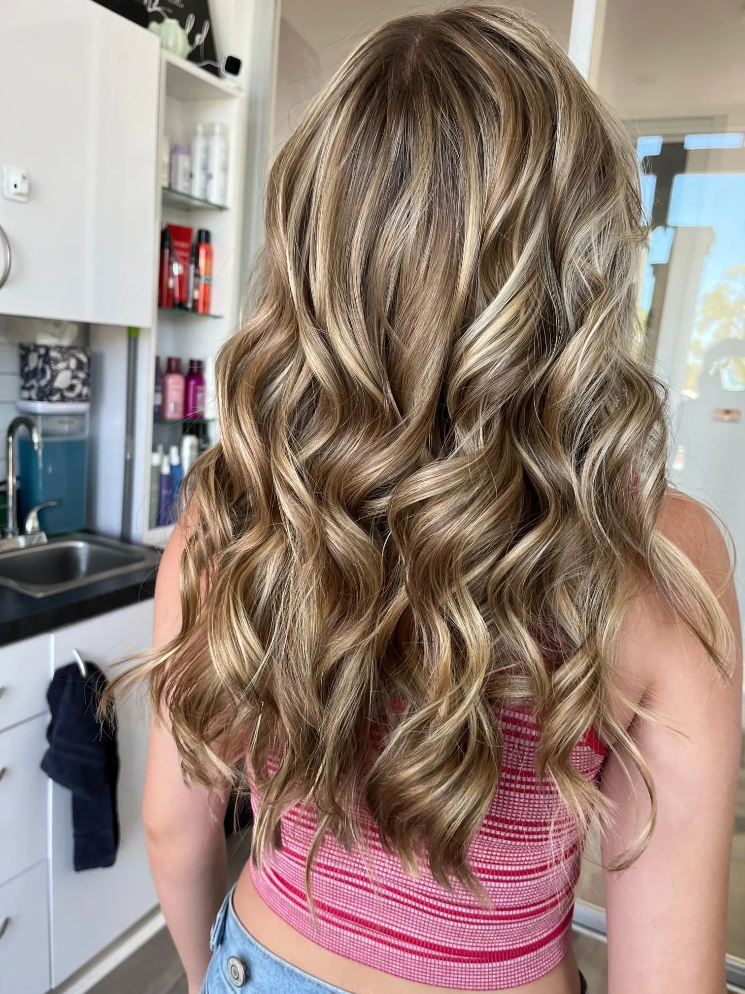 Long blonde balayage hairstyle with soft curls and natural highlights, styled professionally inside a modern hair salon. The look showcases dimensional color blending, smooth waves, and healthy shine.