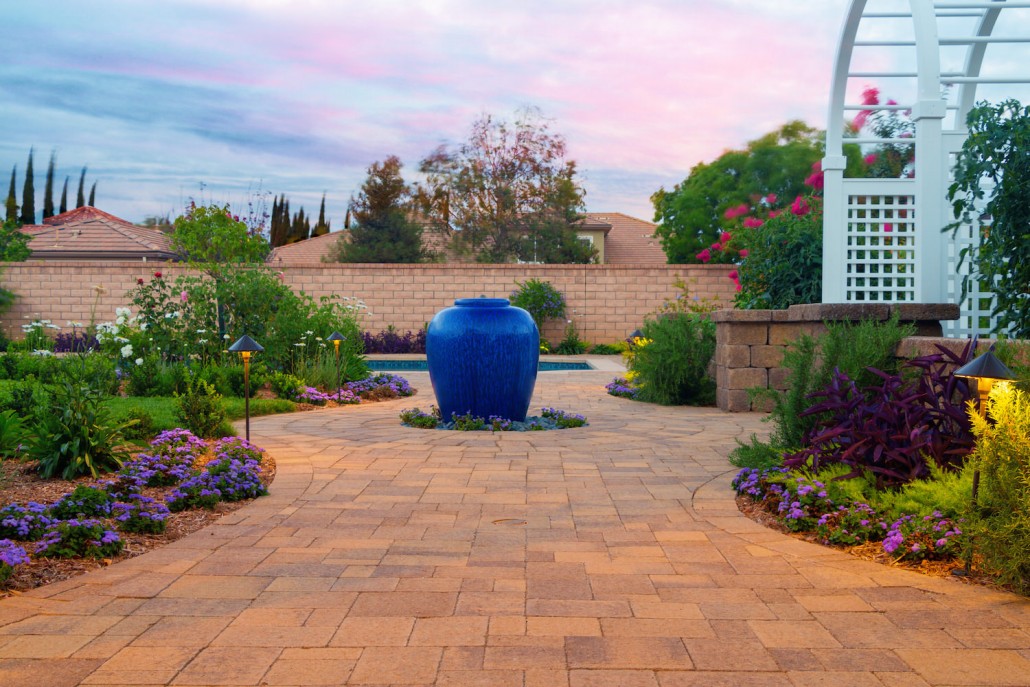 Lush landscaped garden with a bright blue urn on a brick path, colorful flowers, white pergola, and pastel sunset sky creating a serene mood.