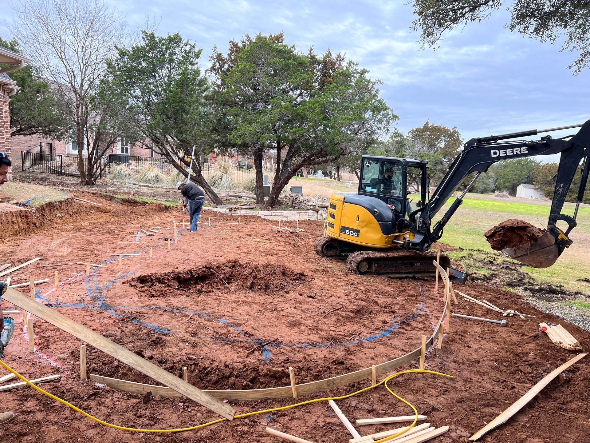 This construction scene shows workers and a John Deere excavator preparing a backyard for a custom pool. The ground is marked with blue paint and wooden forms to define the shape.