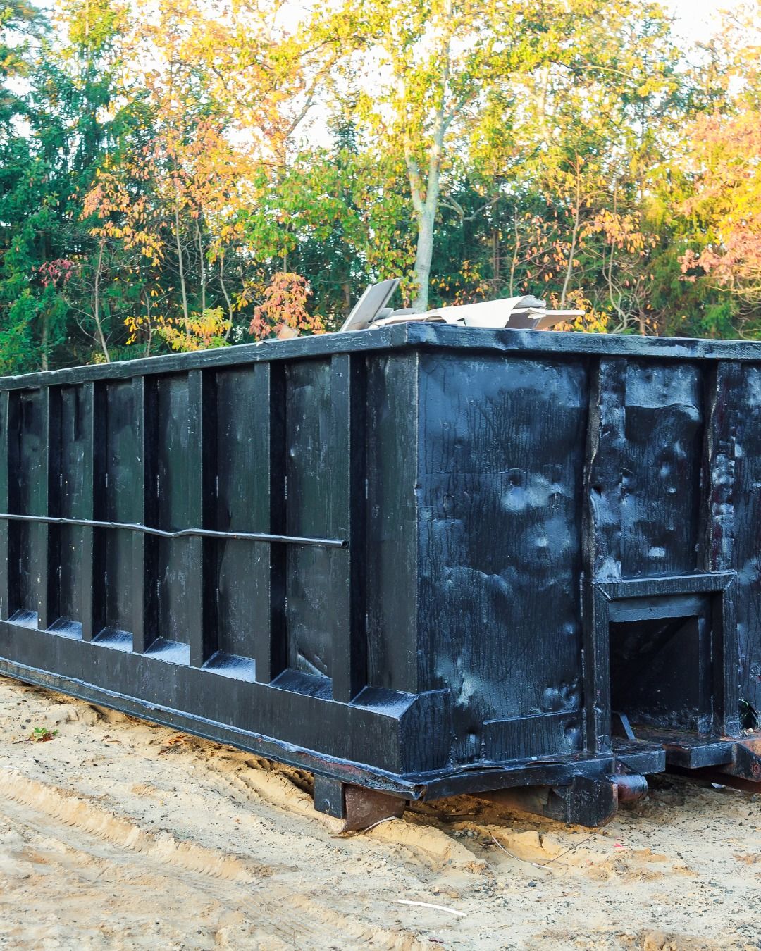 A large black roll off dumpster sits on sandy ground near trees with autumn foliage. Construction debris is visible inside the container, indicating active cleanup or construction work.