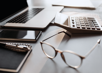Close-up of a professional's desk with laptop, keyboard, cellphone, glasses, and notebook.