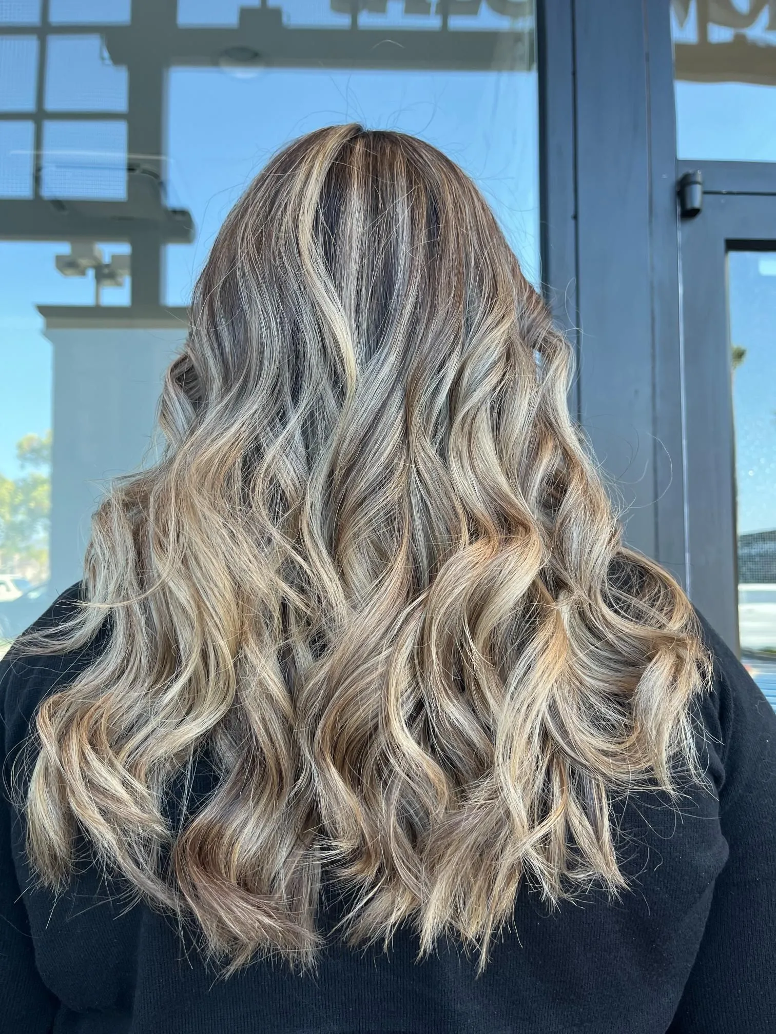 Long wavy blonde balayage hairstyle with soft layered curls and blended highlights, styled professionally to create natural dimension and shine, photographed from the back to showcase color depth.