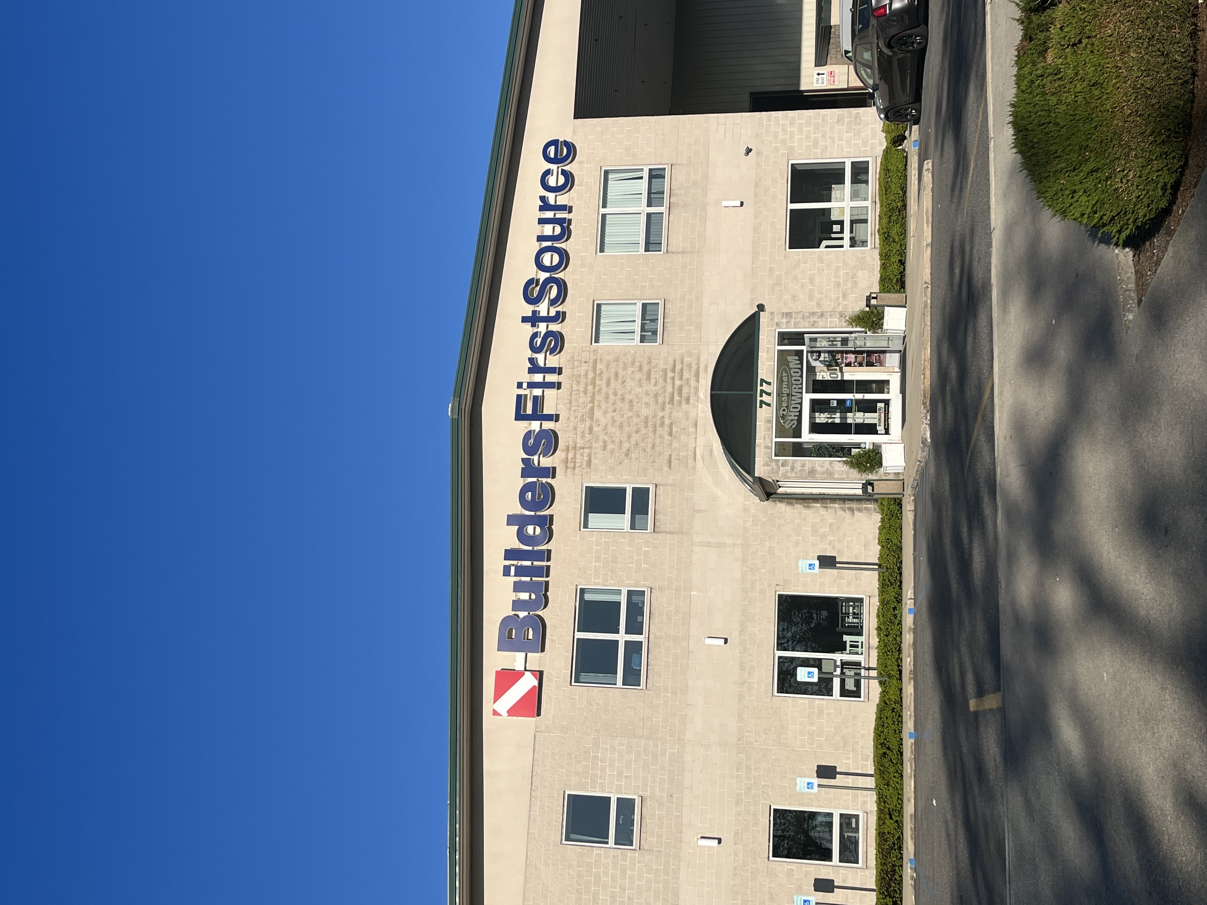 Front exterior of Builders FirstSource building with large company signage, beige brick facade, multiple windows, and a designer showroom entrance with glass doors, set against a clear blue sky and parking lot.