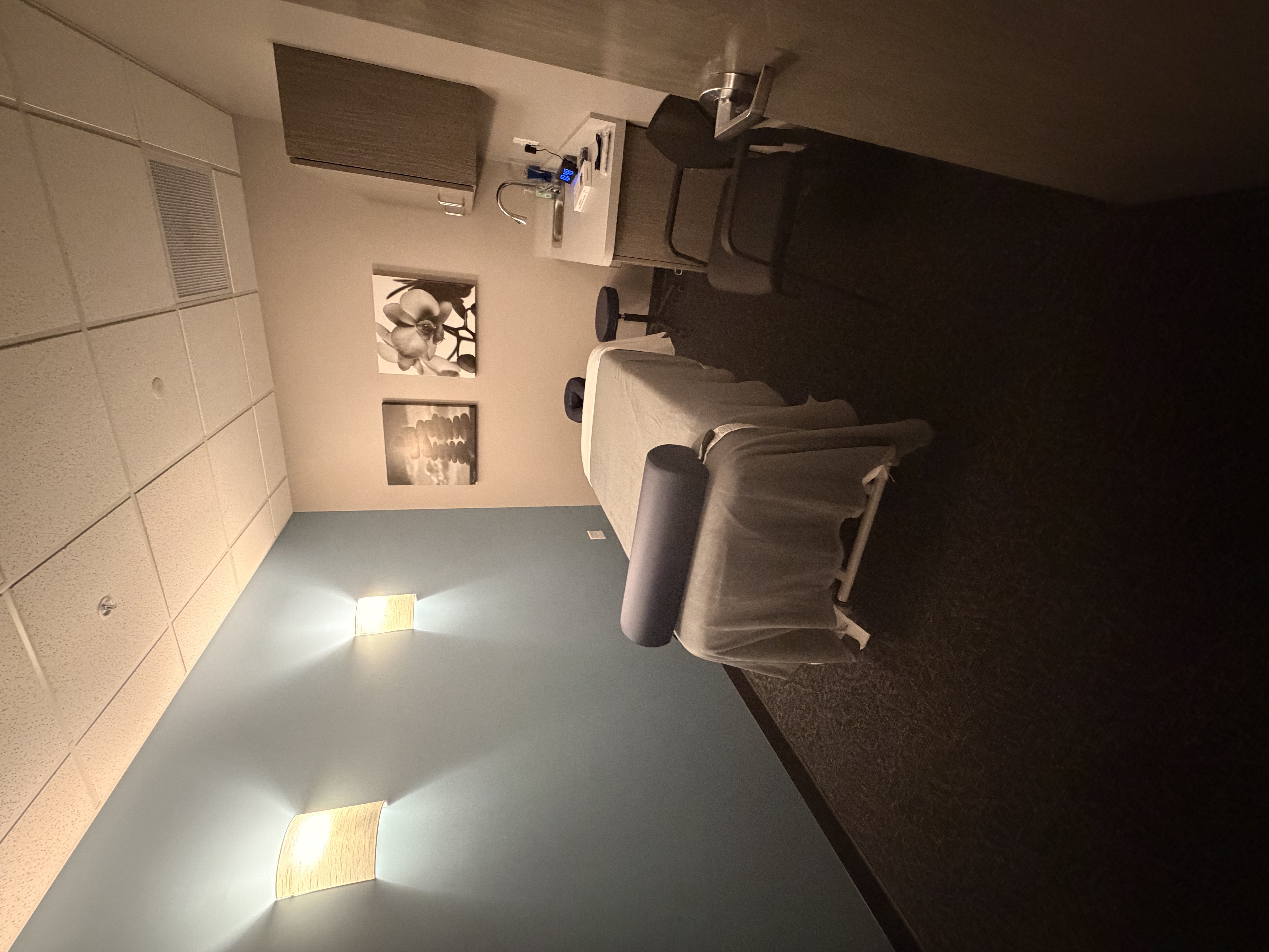 Single massage treatment room with massage table in gray linens. Navy blue bolster and face cradle cover. Gray wood-grain cabinetry with sink and counter. Black and white floral artwork on wall. Soft wall sconce lighting and gray carpet.
