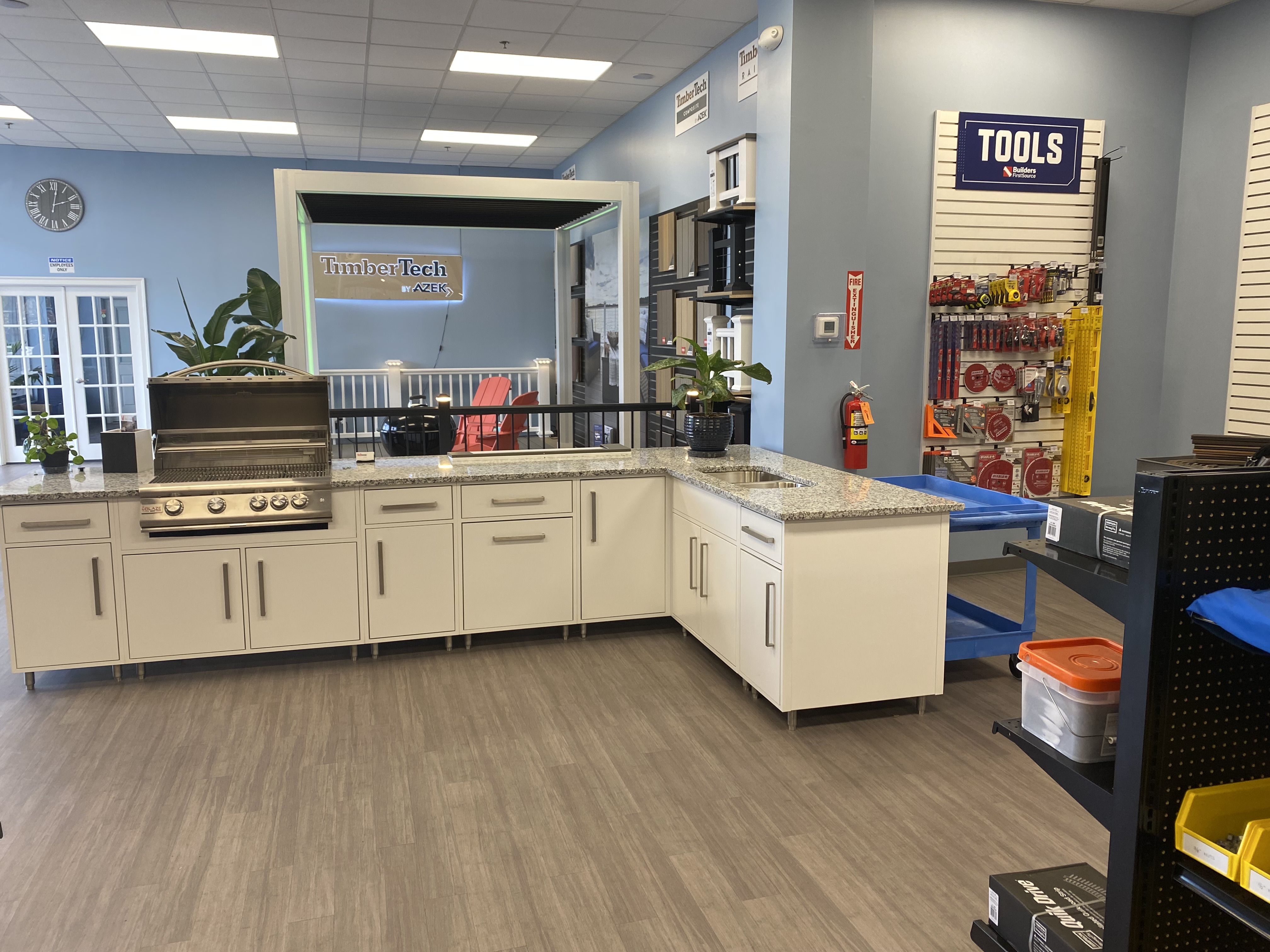 A modern outdoor kitchen display at Builders FirstSource featuring a stainless steel grill, white cabinetry with granite countertops, and a TimberTech pergola. The showroom highlights decking materials, millwork, and tool selection for home improvement and outdoor living projects.